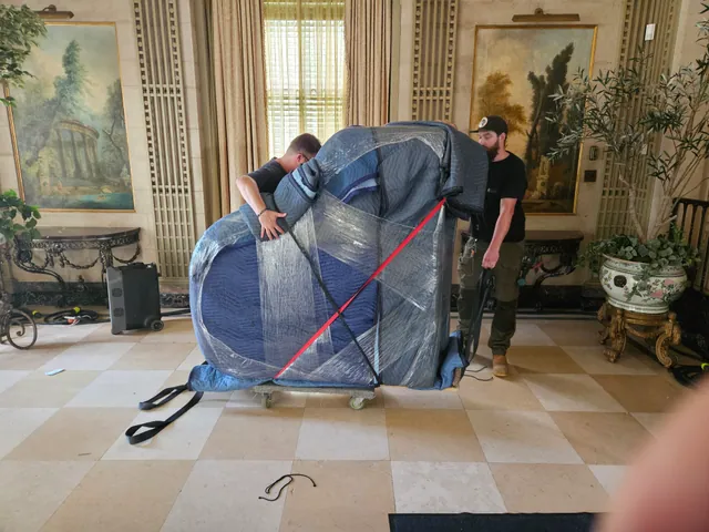 Movers guiding a blanket-wrapped grand piano secured with straps on a dolly